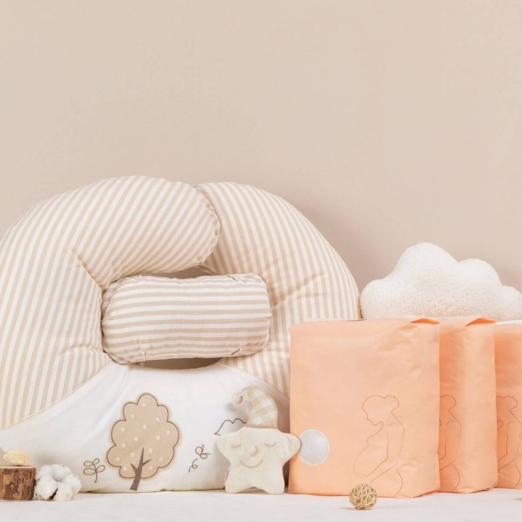 A light pink and white striped nursing pillow is sitting on a while table with a tan star stuffed animal and three peach packages