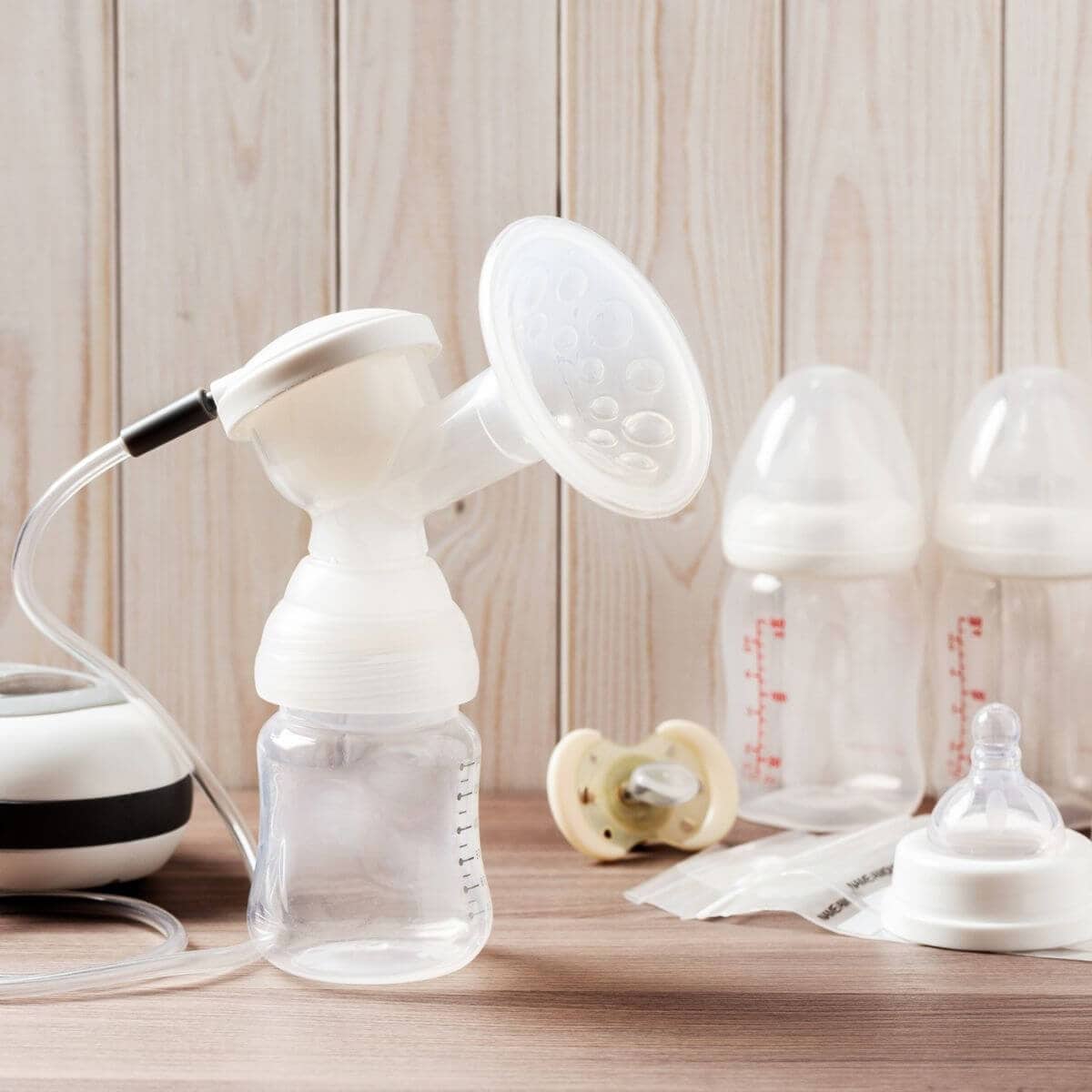A breastpump is sitting on a light wood table with a light yellow pacifier and two baby bottles.