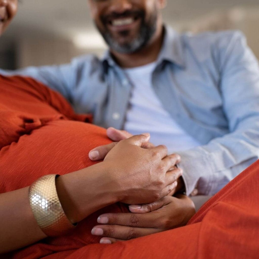 African American couple sitting on a couch with hands on the woman's pregnant stomach