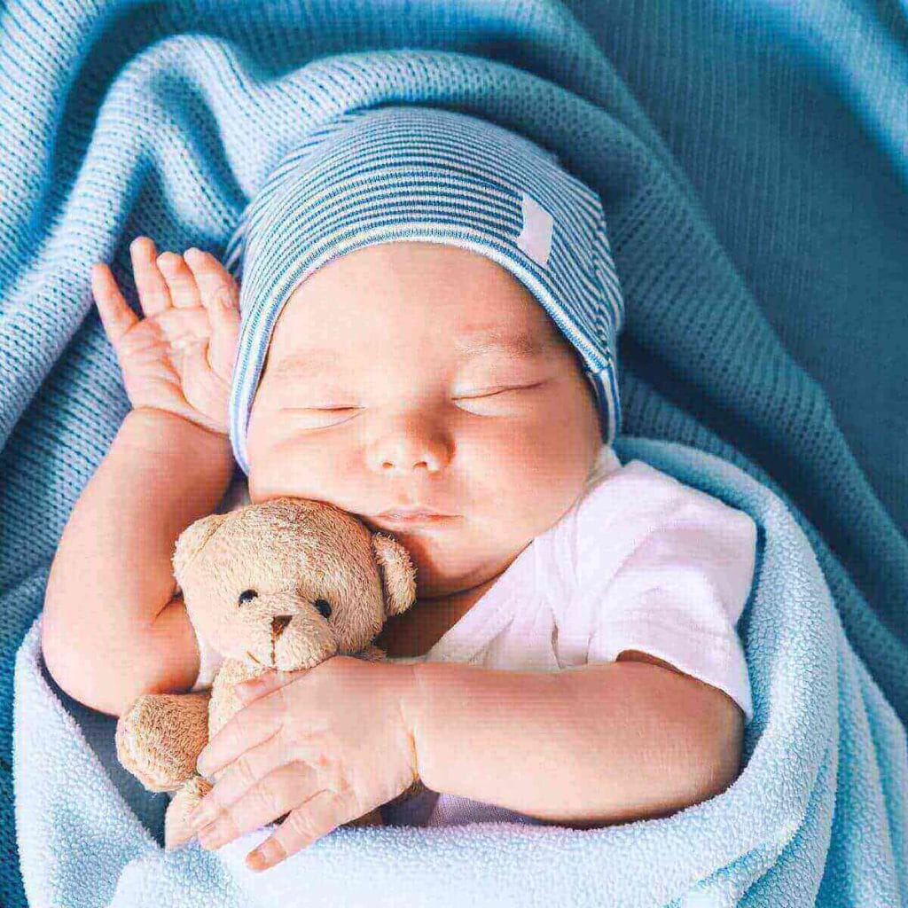 Little boy holding a teddy bear wearing a blue hat and wrapped in blue blanket