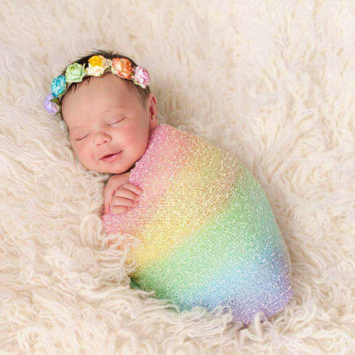 Newborn girl wearing a headband with purple,blue,green,yellow, and orange roses, wrapped in a pink,yellow,green,blue, and purple striped blanket laying on a white shag carpet.