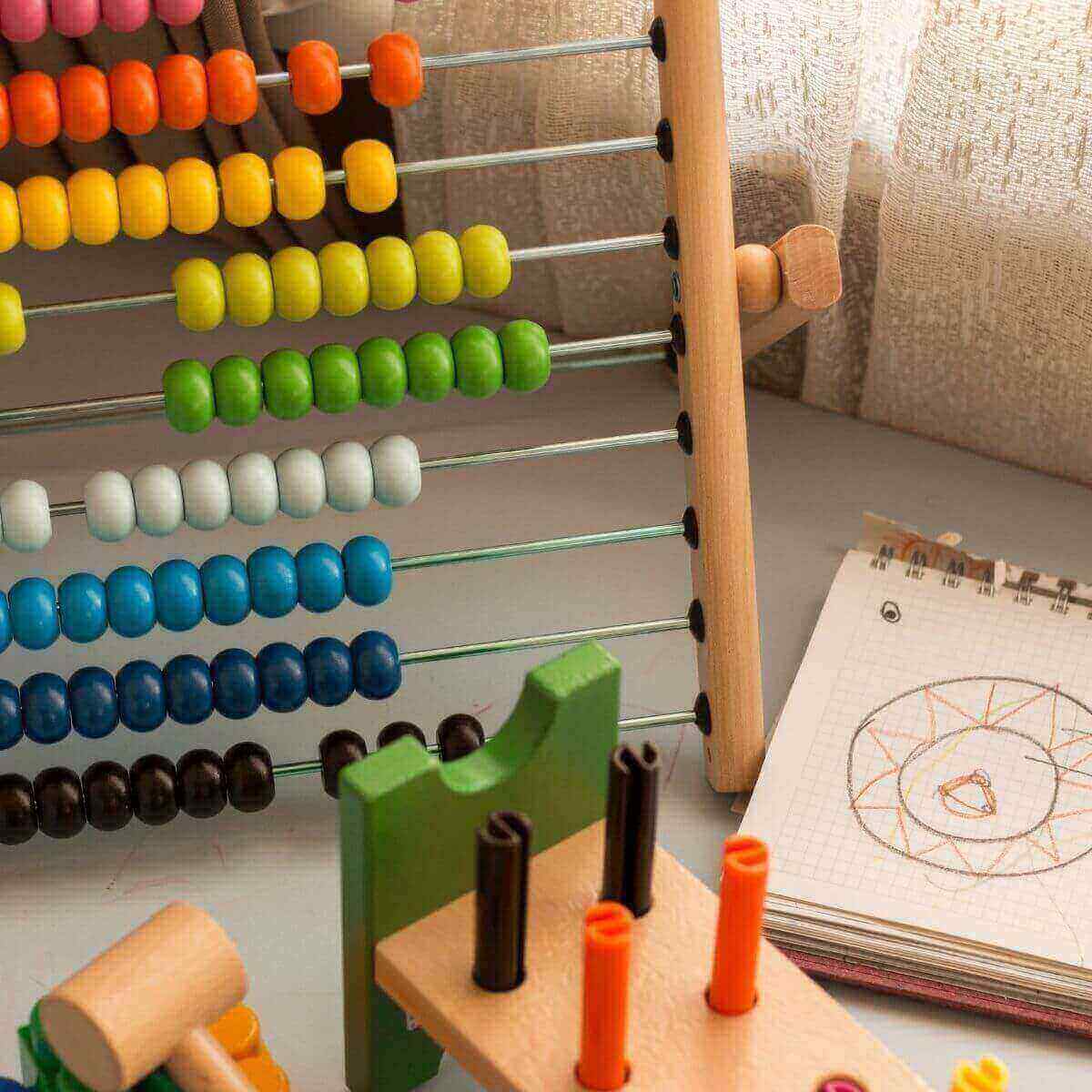 A wooden abacus sits on a table with pink, orange, yellow, green, white, light blue, dark blue, and black beads with a wooden mallet and a peg board.