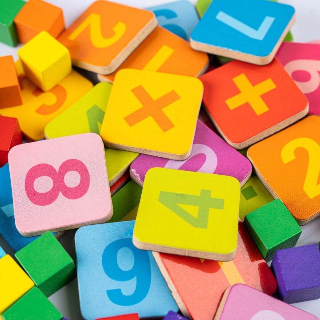 Wooden squares that have numbers on them are piled up in the middle of a table. The squares are light pink, yellow, purple, blue, red, and orange.