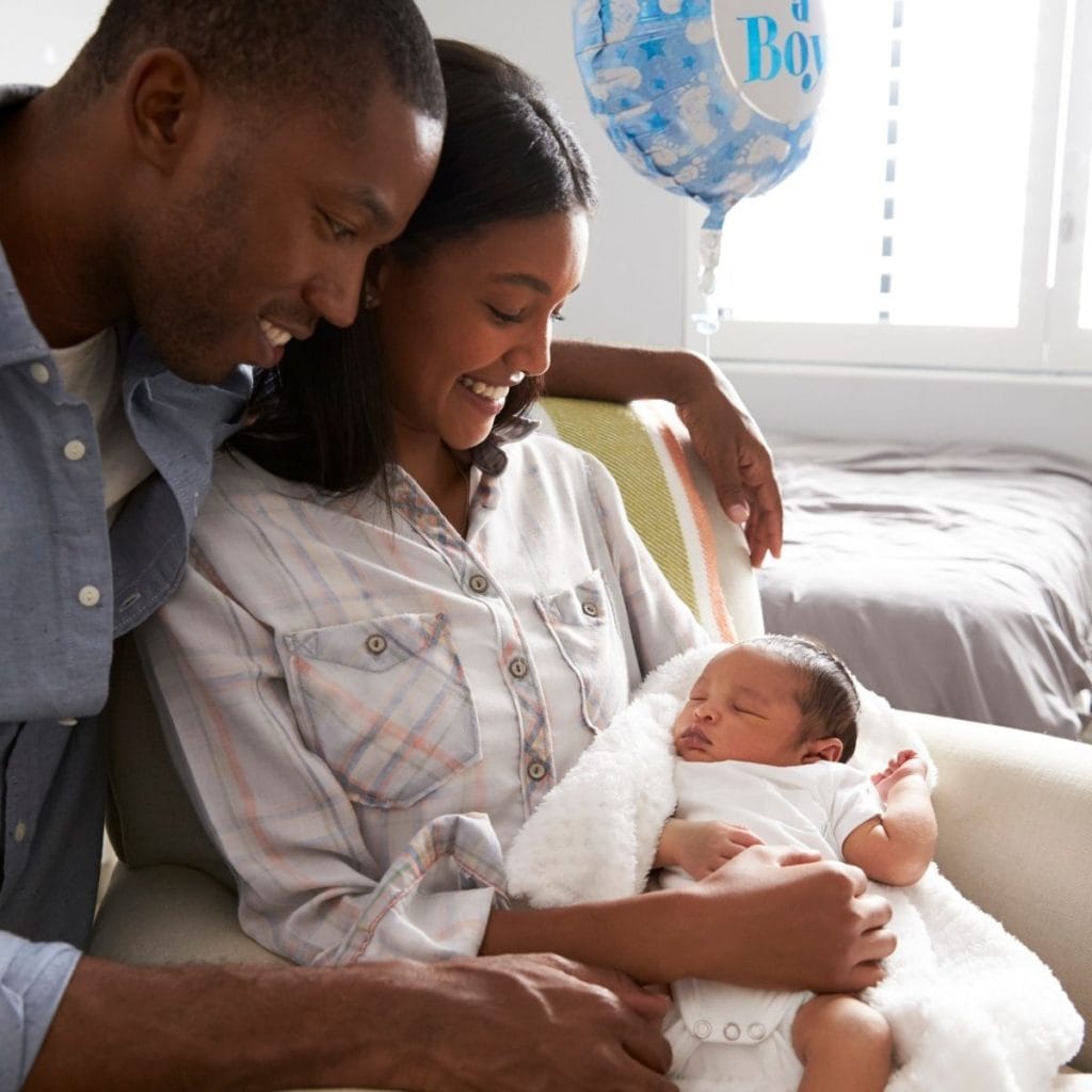 African American couple is in a bedroom. The male is wearing a white t-shirt with a denim long sleeve shirt over it. The woman is wearing a orange and grey plaid shirt while sitting in a chair. Both are smiling down at the newborn in the woman's lap.