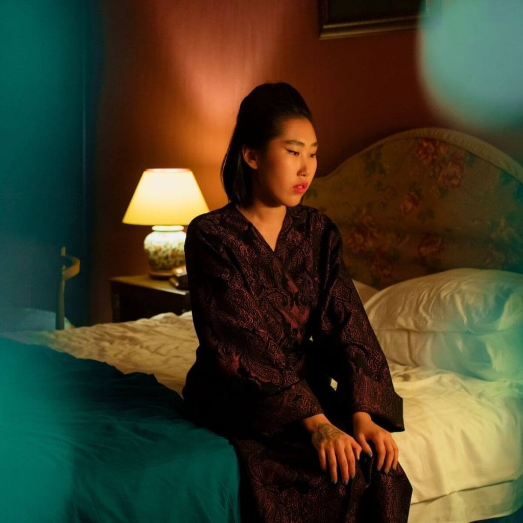 An Asian American woman is sitting on the side of her bed, wearing a dark purple robe with a black paisley design on it, staring down at the floor and thinking.