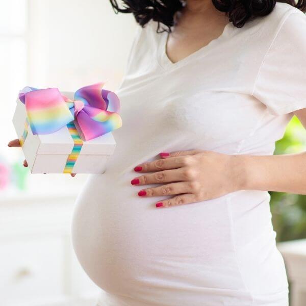 A pregnant woman with shoulder length black hair is wearing a white teeshirt. Her left hand is on her belly and she has a white gift box in her right hand with a rainbow colored bow.