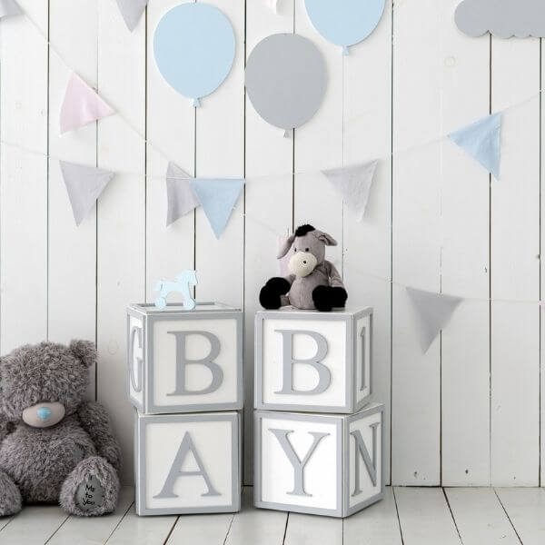 A white wooden slatted wall has triangle banners and balloons on it in grey, light pink, and light blue. There is a teddy bear in the bottom left corner on the wooden slatted floor with a light blue nose. White and grey blocks spell out the word Baby.