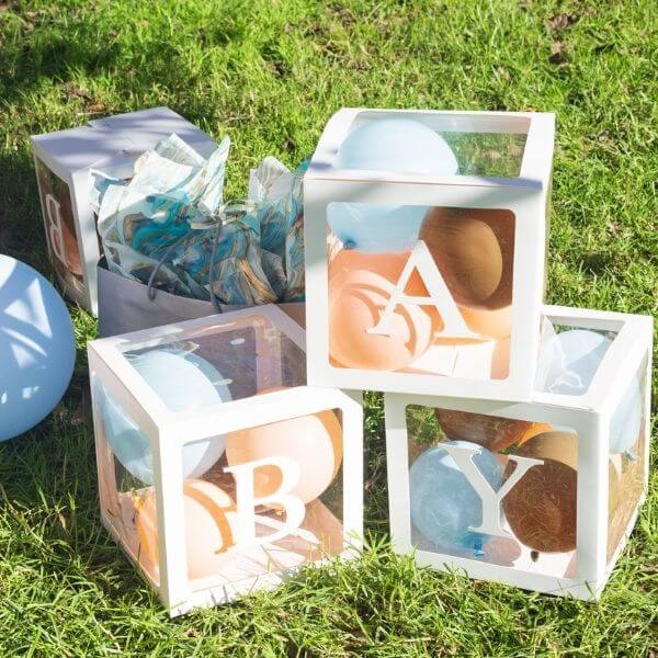 Clear blocks with the letters "B", "A", and "Y are stacked to spell baby. They are sitting on the grass with a blue balloon on one side and a grey gift bag with silver tissue paper coming out of the top