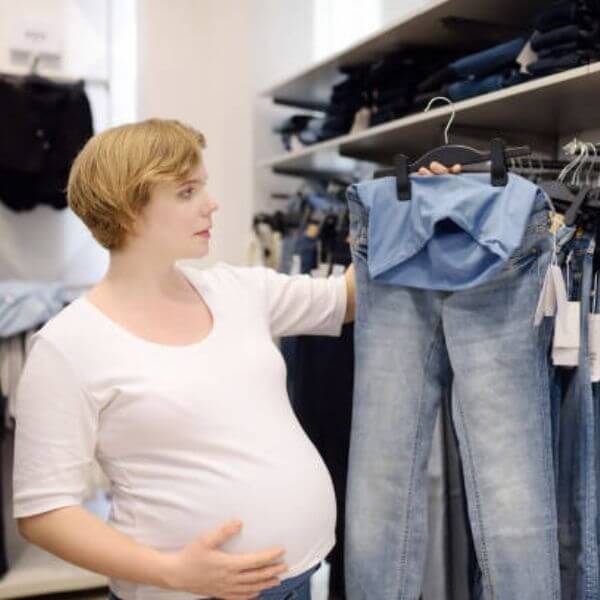 A pregnant woman with her hand on her belly is standing in a store holding up a pair of light blue maternity jeans