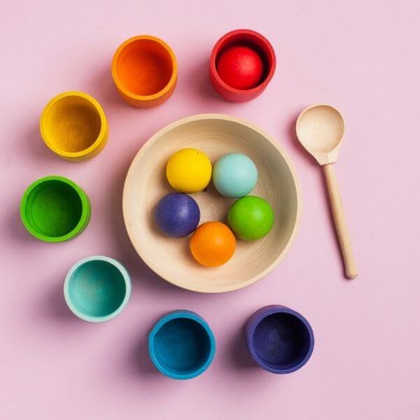 A light wood bowl is sitting in the middle of a light pink table. In it there are yellow, light blue, green, orange, and dark blue wooden balls. Around the wooden bowl are a wooden spoon and wooden bowls that are red, orange, yellow, green, light blue, dark blue, and purple.