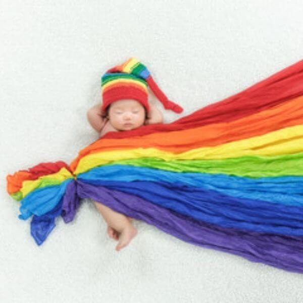 An Asian American baby is laying on a white blanket. The baby is wearing a long stocking cap that has red, orange, yellow, green, and blue stripes. The baby is covered with a material that has red,orange,yellow,green,light blue, dark blue, and violet.