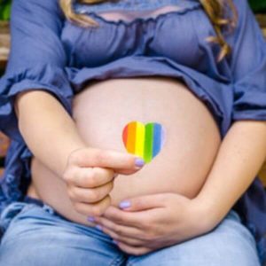 A woman is sitting on a wooden bench. She is wearing a dark blue top and light blue jeans. She has one hand on her pregnant belly and the other hand is holding a heart with red, orange, yellow, green, blue, and violet stripes.