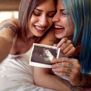 Two women are laying on a bed on their stomachs. One has blue hair and the other has dark brown hair. They both have tattoos on their arms and are holding a ultrasound picture while smiling.