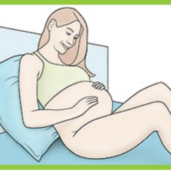 Woman lies on bed propped on pillow with her knees bent and her hands on her pregnant belly