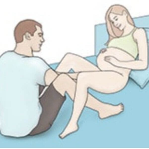Pregnant woman sits on bed, reclined against a pillow, with her legs gently bent. A man sits across from her with his legs also bent next to her legs. 