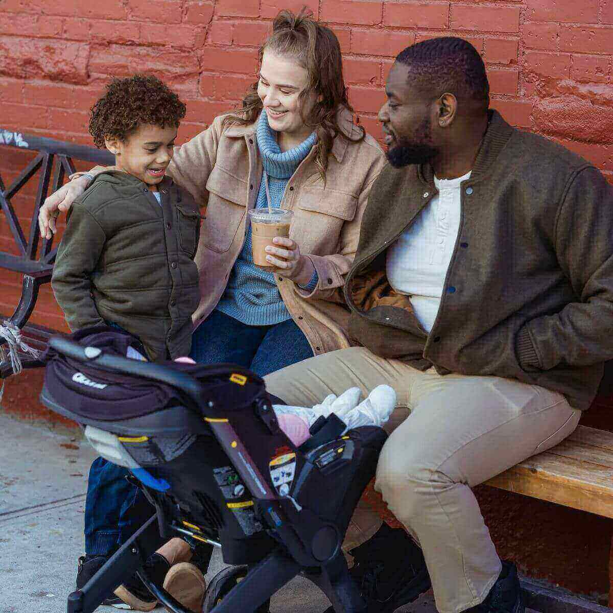 An African American man and a Caucasian woman are sitting on a bench with an older biracial boy and a newborn in a stroller in front of them