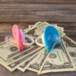 Pink and blue pacifiers are sitting on top of two twenty dollar bills, two two dollar bills, and two one dollar bills. These are sitting on top of a wooden table.
