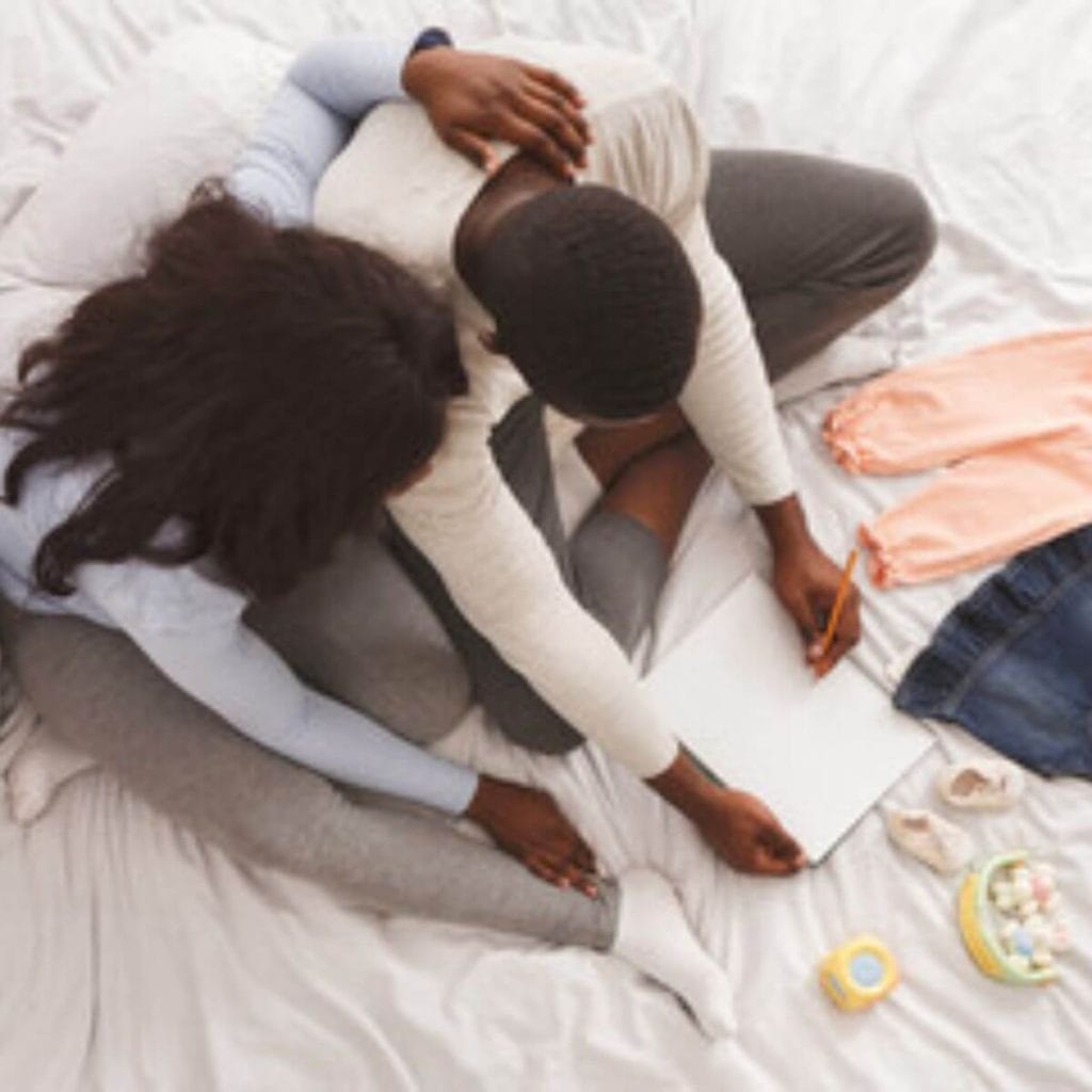 An African American couple is sitting on a bed with a white sheet. There are a pair of peach baby pants, a dark denim baby dress and baby booties laying in front of them. The man has a notebook and pencil.