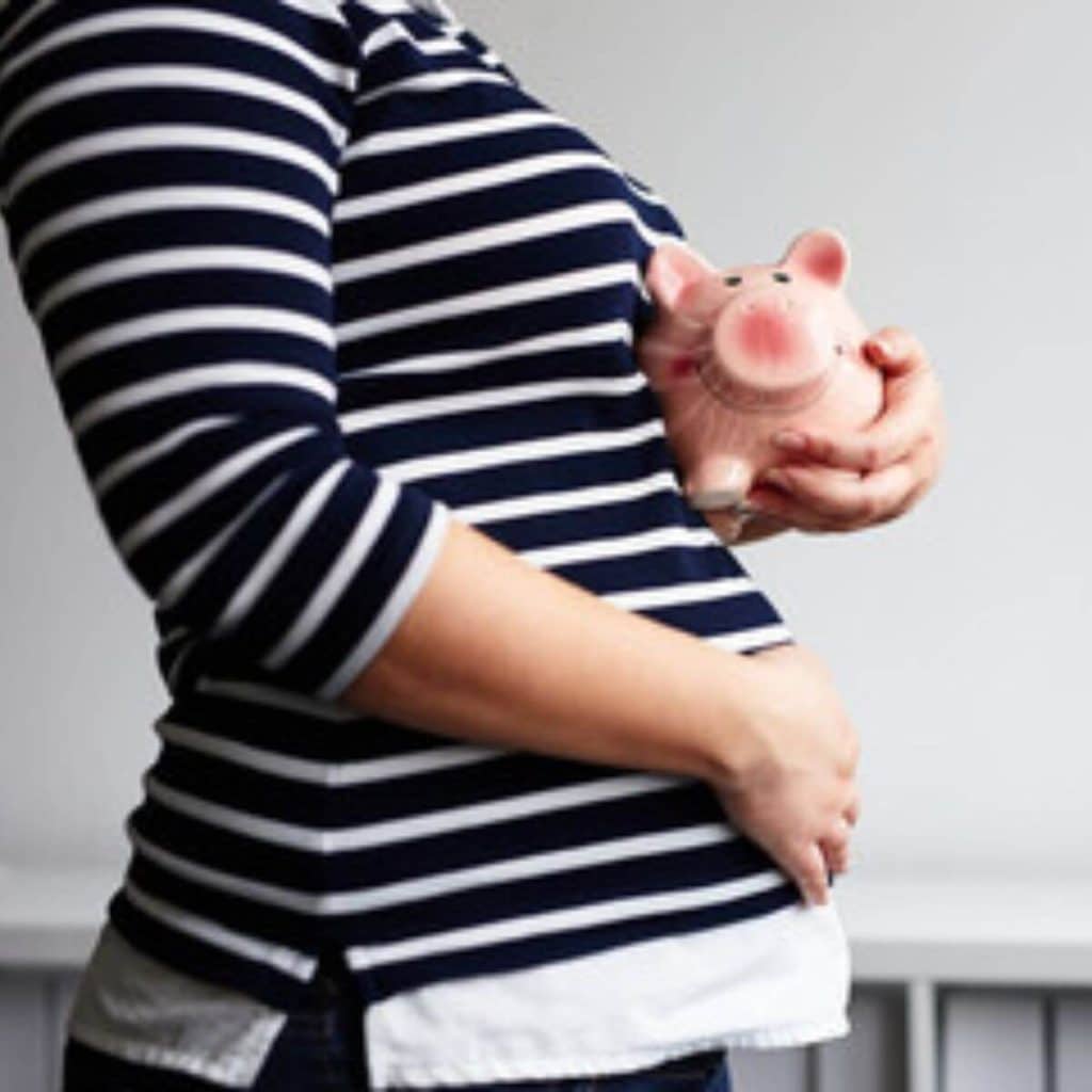 A light grey wall is in the background. A pregnant woman with a black and white striped shirt has her right hand on her stomach and is holding a pink piggy bank in her left hand.
