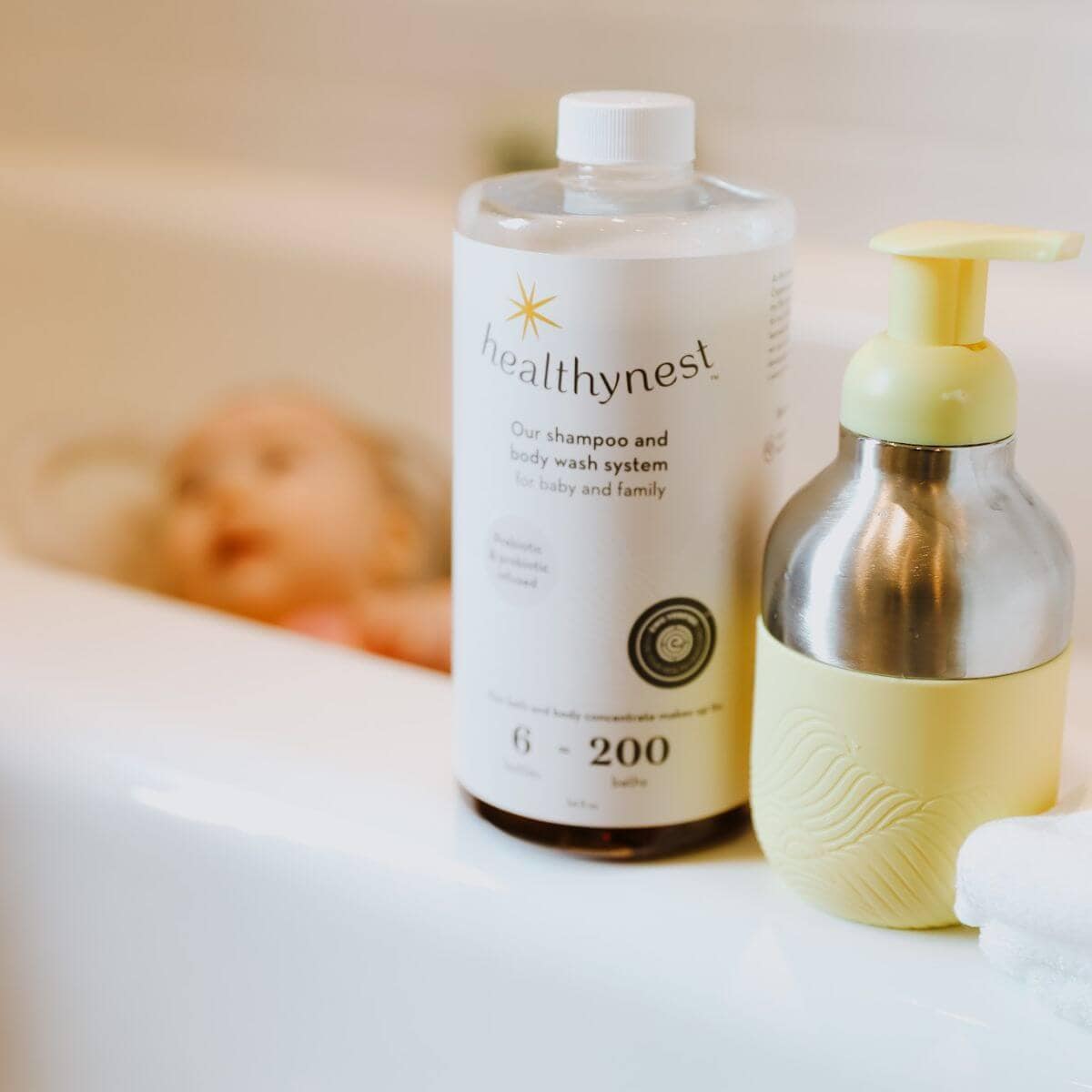 A baby is in a baby tub. On the side of the tub is healthybaby shampoo and a pump bottle that is silver and pale yellow