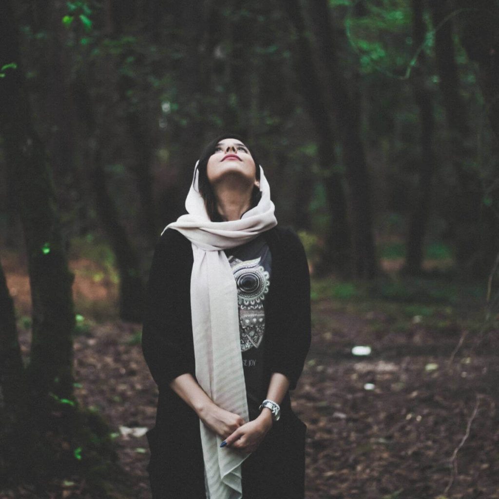 A woman is standing in the middle of the woods. She has on a floor length black sweater and a white scarf wrapped around her head. She is looking up to the sky with her hands in front of her.