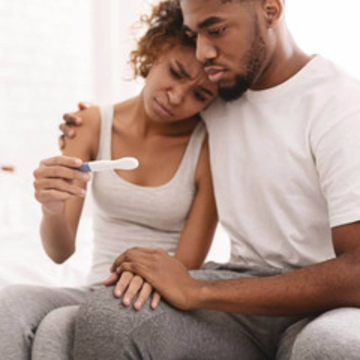 An African American man and woman are sitting on the edge of a bed. She is wearing a white tank top and grey pants and he is wearing a white teeshirt and dark grey sweats. They are looking down at a pregnancy test with confused looks on their faces.