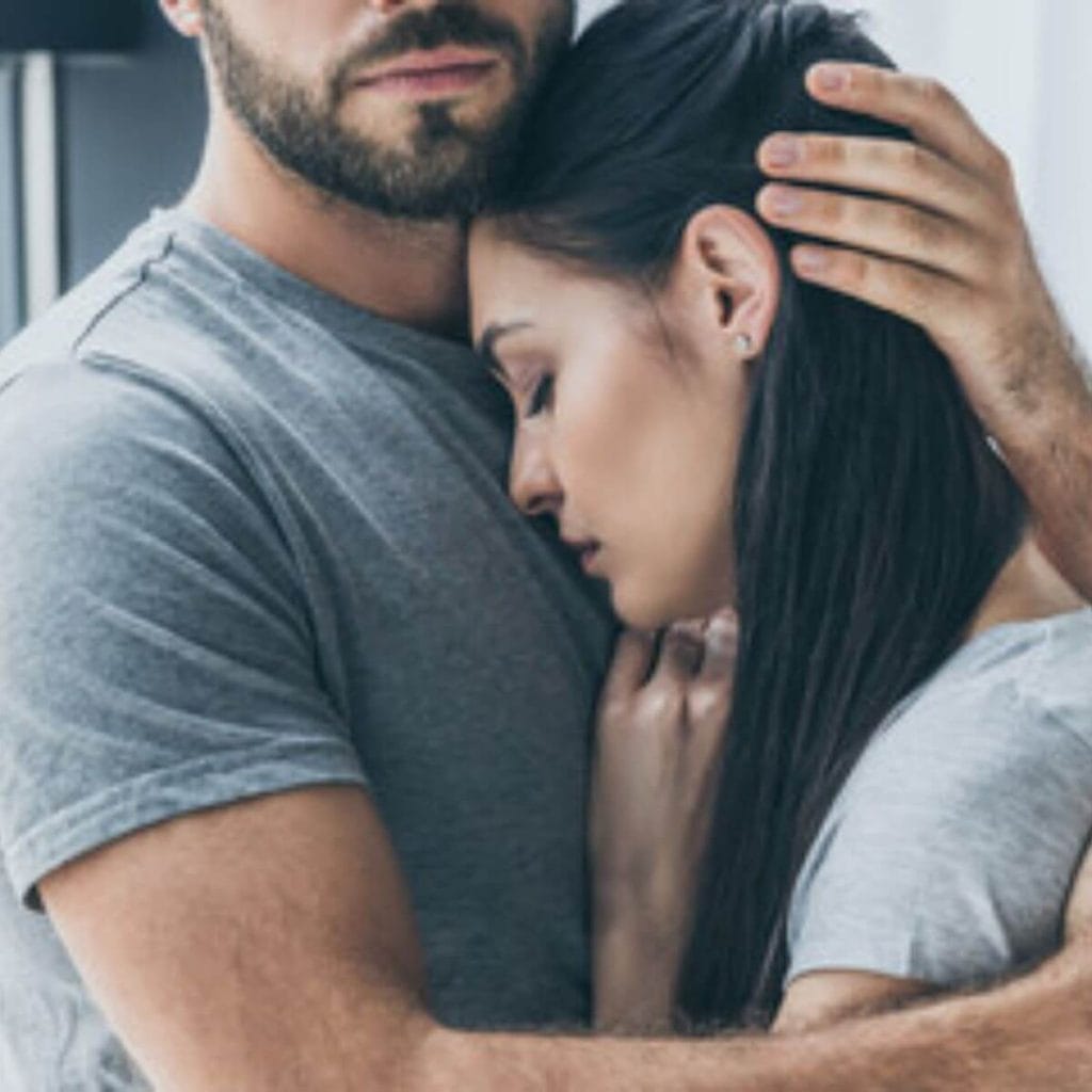 A Caucasian couple is standing up. The man has a beard and mustache and is wearing a dark grey tee-shirt. He is hugging a woman with long black hair with a light grey tees-shirt who has her eyes closed and looks sad.