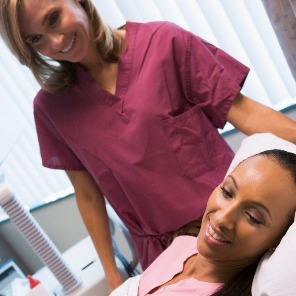 An African American woman wearing a light pink top is laying on an examination table with a caucasian nurse wearing rose colored scrubs standing beside her.