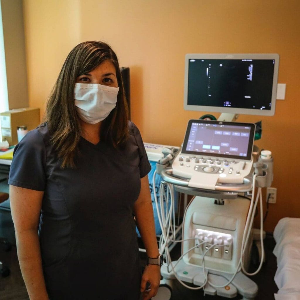A woman with long dark brown hair is wearing navy blue scrubs. She is standing in front of an ultrasound machine in a doctor's office room.