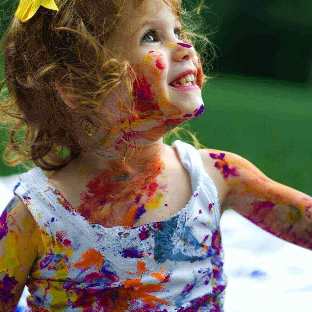 A little girl with a yellow bow in her hair is looking to her left. She is wearing a white tank top and is covered in red, yellow, purple, orange, and blue paint.