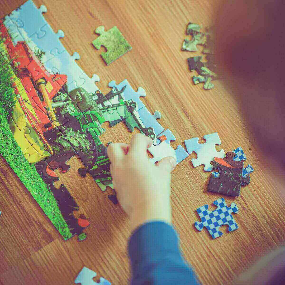 The hand of a toddler is putting together a puzzle on a wood floor with a red and green tractor on it.