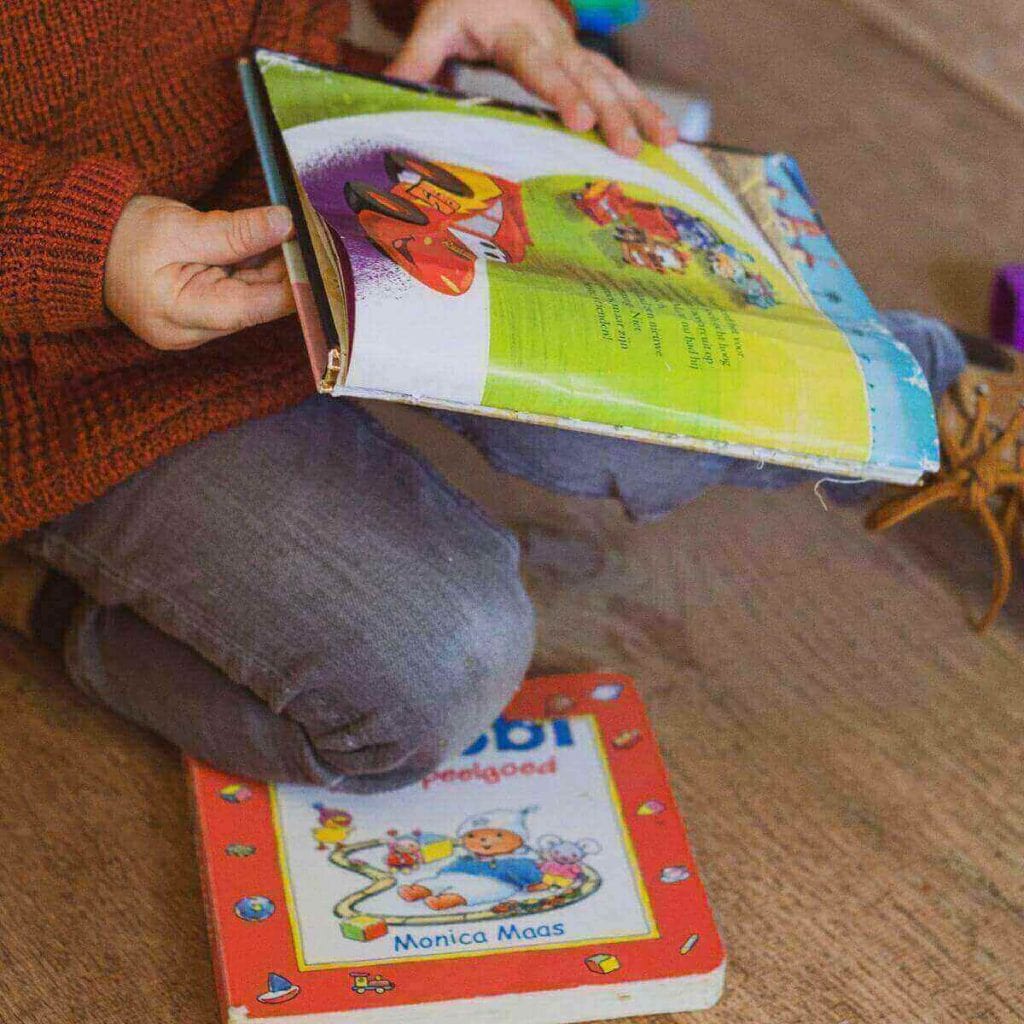 A boy wearing an orange sweater and grey pants is sitting on a wooden floor. In his hands, he has a book and another book is under is knee.