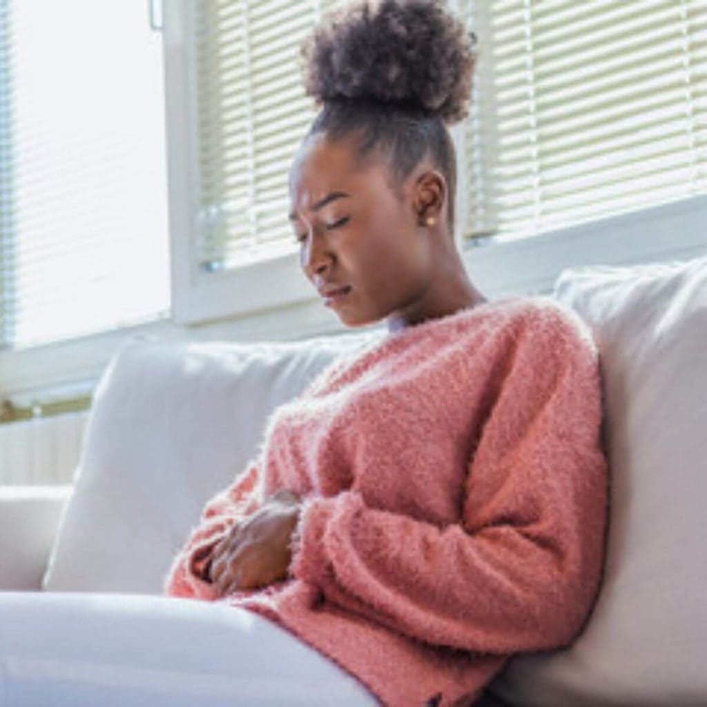 An African American woman is sitting on a cream colored couch. Her eyes are closed in pain and she has her hands on her stomach.