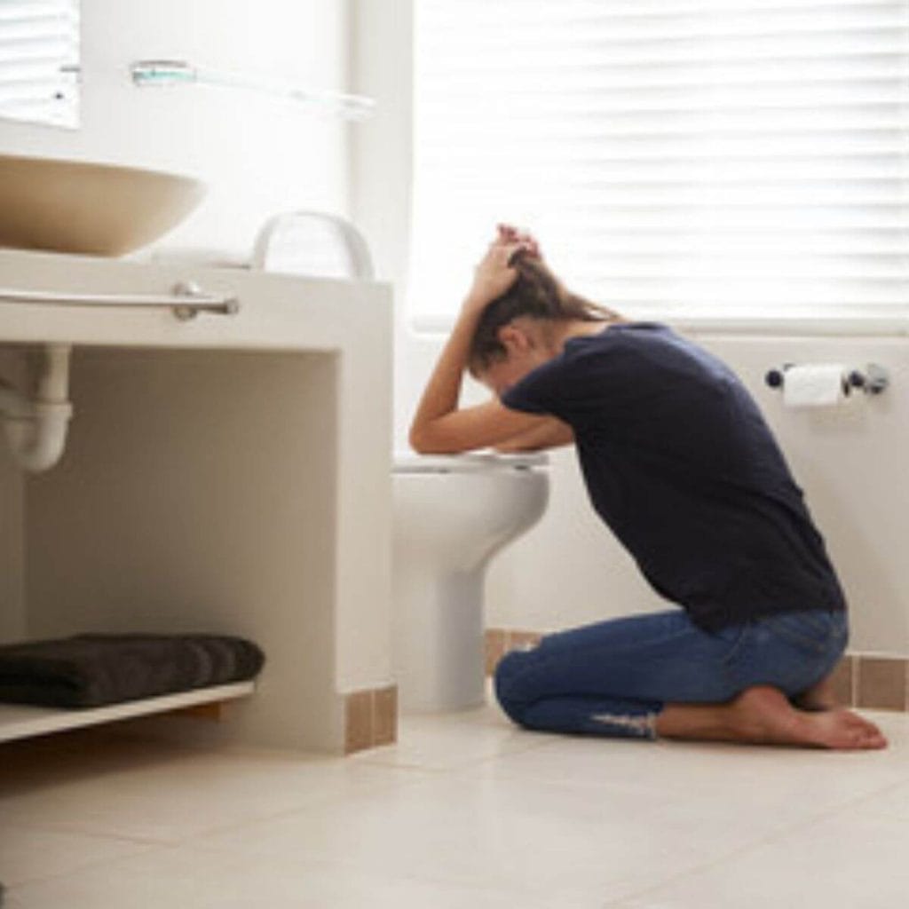 A woman wearing a black tee-shirt and dark colored jeans is in an all white bathroom leaning over the toilet with her hands on the sides of her head.