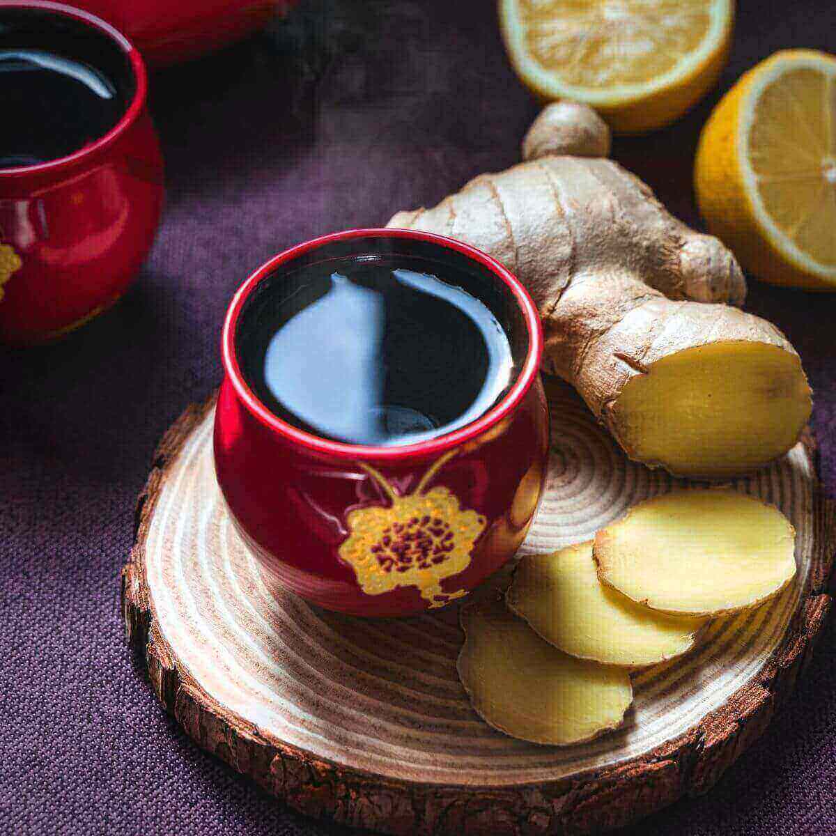 A red cup of tea is sitting on a round piece of wood. There are slices of ginger beside the cup with a cut lemon in the background
