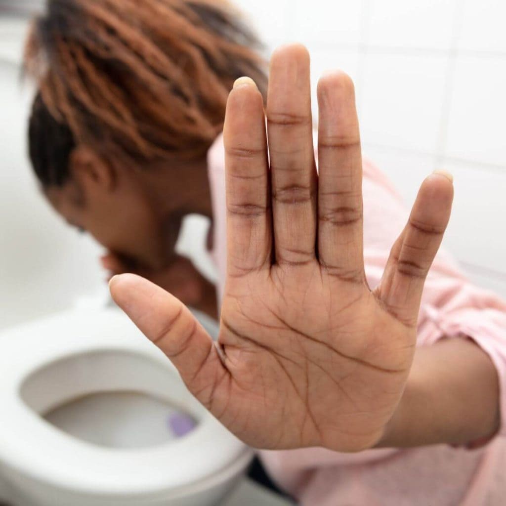 An African American woman is in an all white bathroom. She is wearing a light pink shirt with her hair in a ponytail. She has her left hand up in the stop gesture and she is leaning over the toilet.