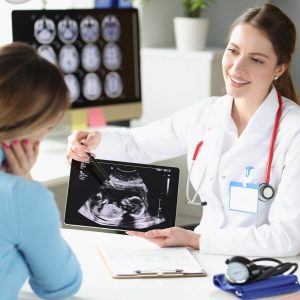 An obstetrician is sitting at a table across from a woman who is wearing a light blue sweater with her right hand on the side of her face. The doctor is showing the woman an ultrasound picture on a tablet.