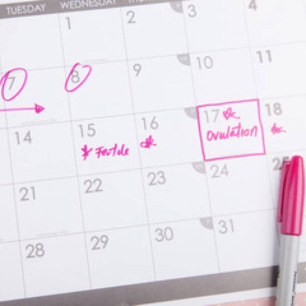 A white and grey calendar is sitting on a light pink table. A hot pink marker is lying off to the side. The 7th and 8th are circled in hot pink and the 17th is blocked off with the word "Ovulation"