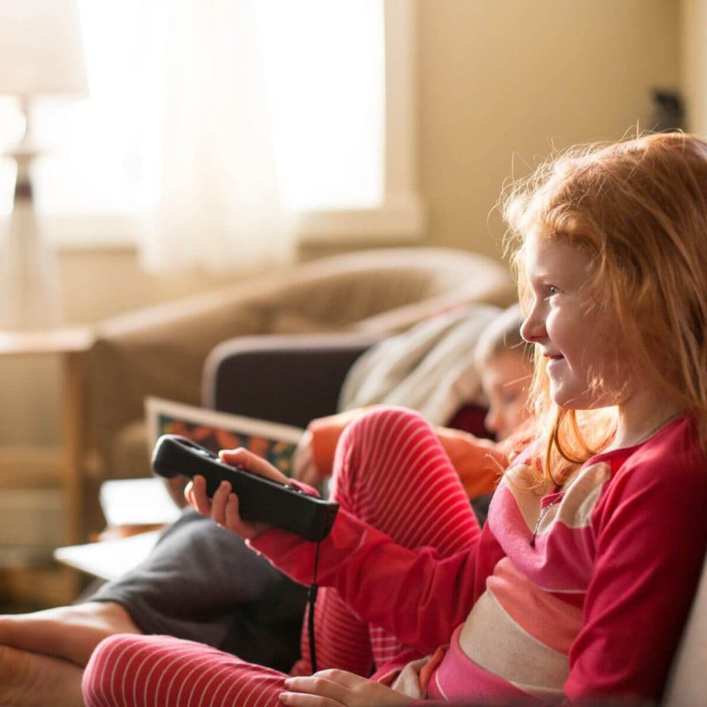 A red headed girl in pink and white striped pajamas is sitting on a couch with a boy in the background. She is holding a television remote and smiling