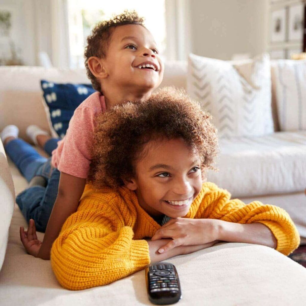 An African American girl wearing a yellow sweater and jeans in laying on her stomach smiling at the television. An African American boy wearing a red short sleeve shirt and jeans is sitting on her back smiling at the television.