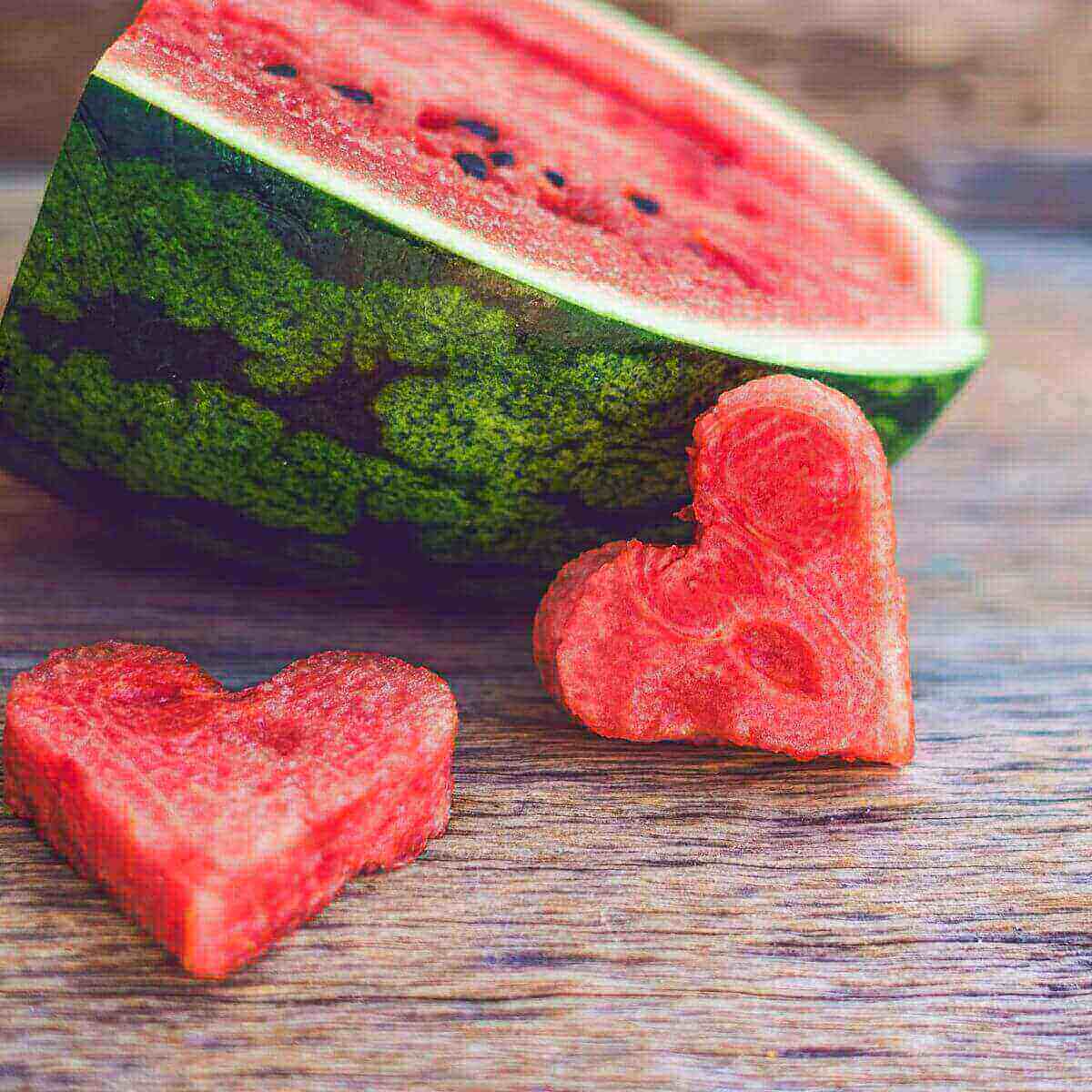 On a wood grained table sits one quarter of a watermelon. In front is two hearts cut from the watermelon.