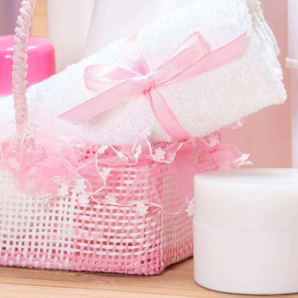 On a light wood table sits a white jar of cream, a pink basket, and a white towel wrapped with a pink bow
