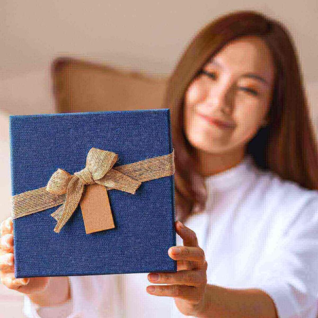 A woman with medium length dark brown hair wearing a white shirt is smiling and holding out a dark blue box with a bow on it.