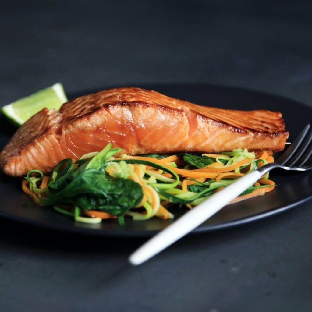 On a dark grey table sits a black plate with a white handled fork. On the plate is a piece of salmon on top of a bed of leafy greens, shredded zucchini, and shredded carrots