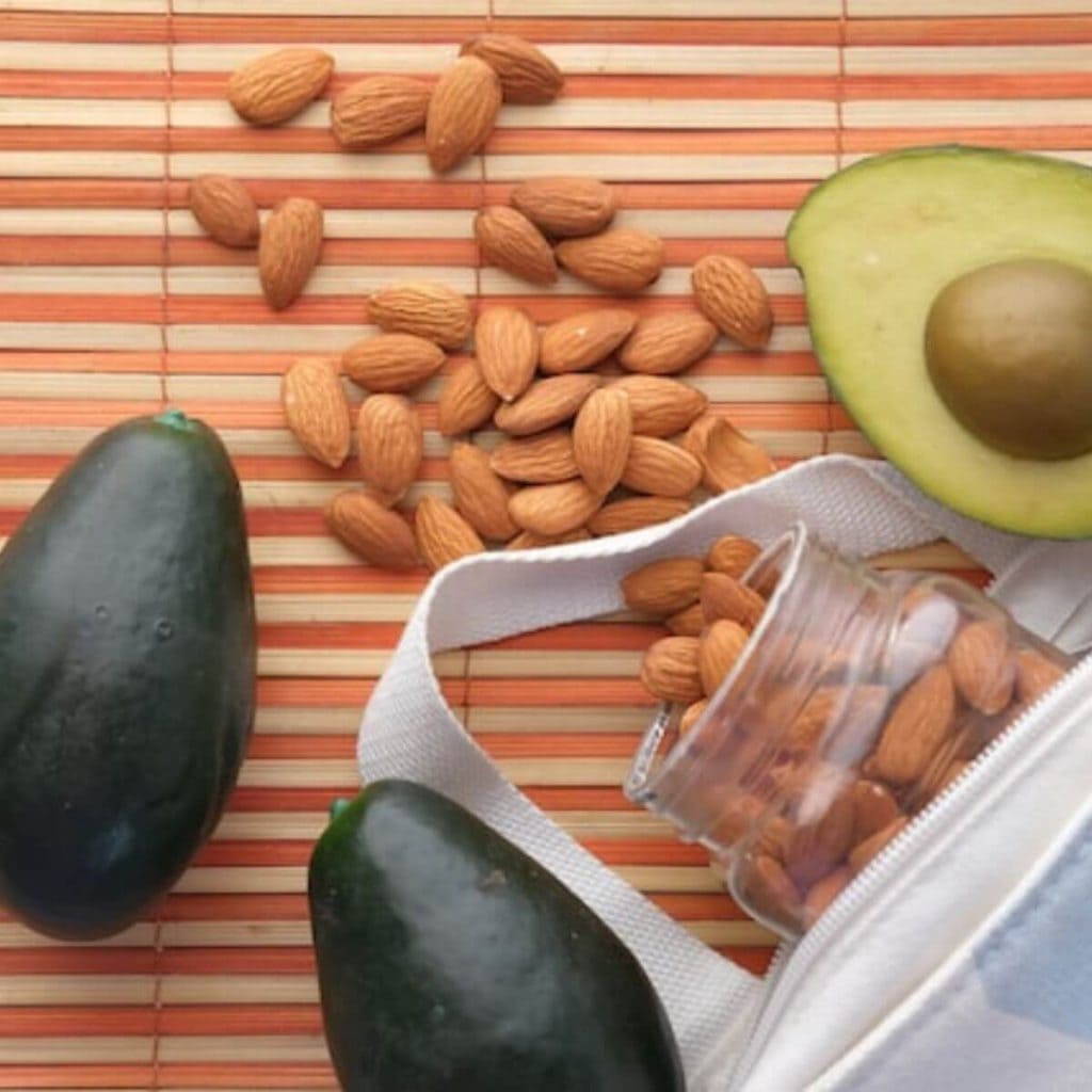 On a cream and orange stripped mat there is a white bag with a clear jar of nuts scattered around and 3 avocados.