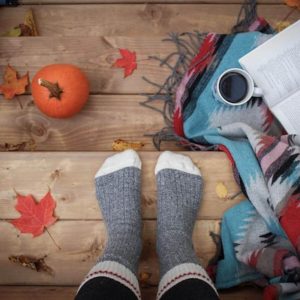 A woman's feet are in grey and white comfy socks. Her feet are on wooden steps with a cup of coffee, a blanket, and a book with orange, yellow, and red leaves.