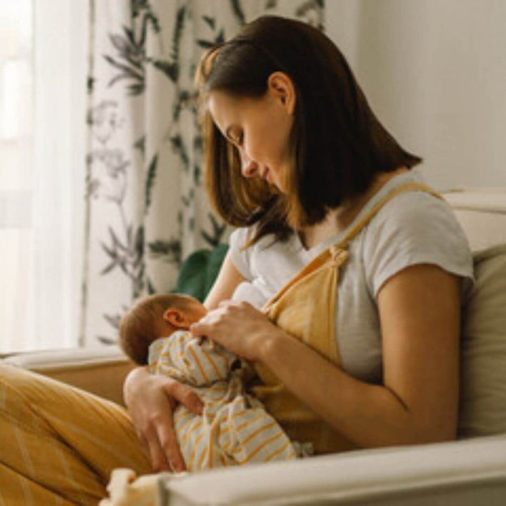 A woman is sitting in a cream colored chair. She is wearing mustard colored overalls and a white shirt. She is nursing her newborn who is wearing mustard and white striped footie pajamas