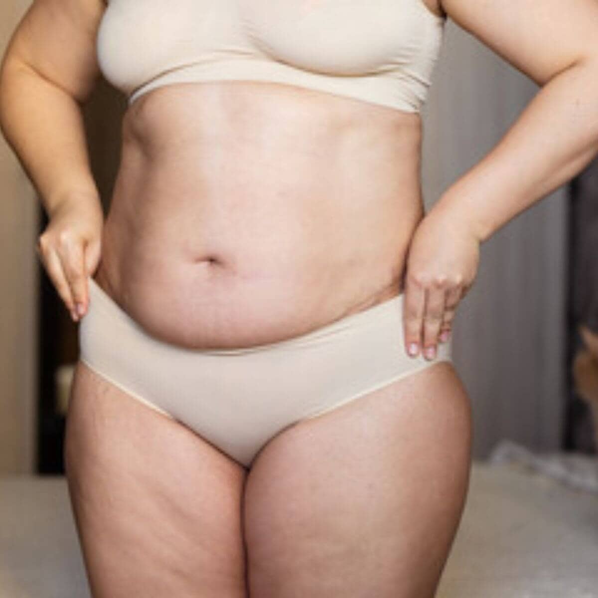 A woman is standing in her bedroom wearing a tan colored bra and panty set with her hands on the waist of the panties