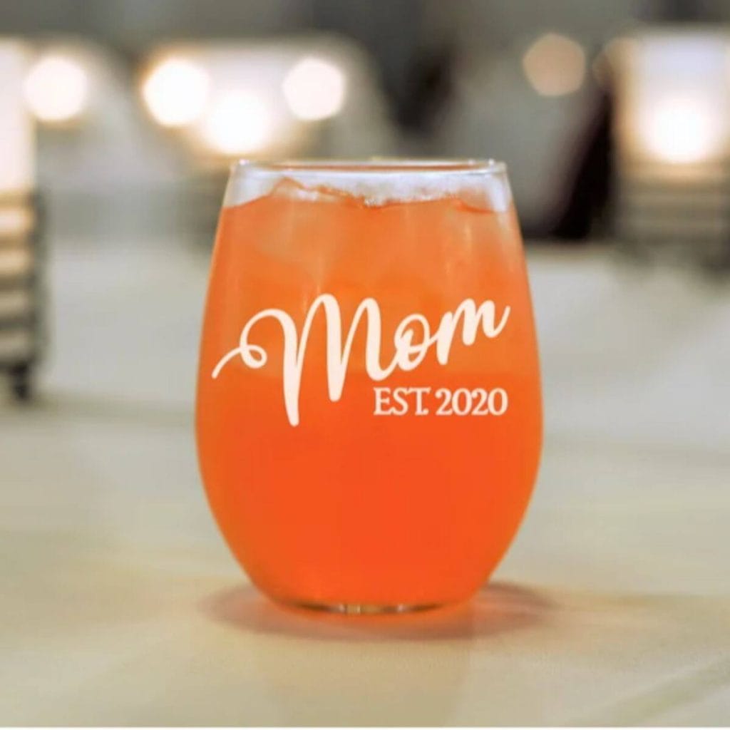 A stemless wine glass is sitting on a marble countertop. The glass says "Mom EST. 2020" and there is a pinkish orange liquid with ice in it.