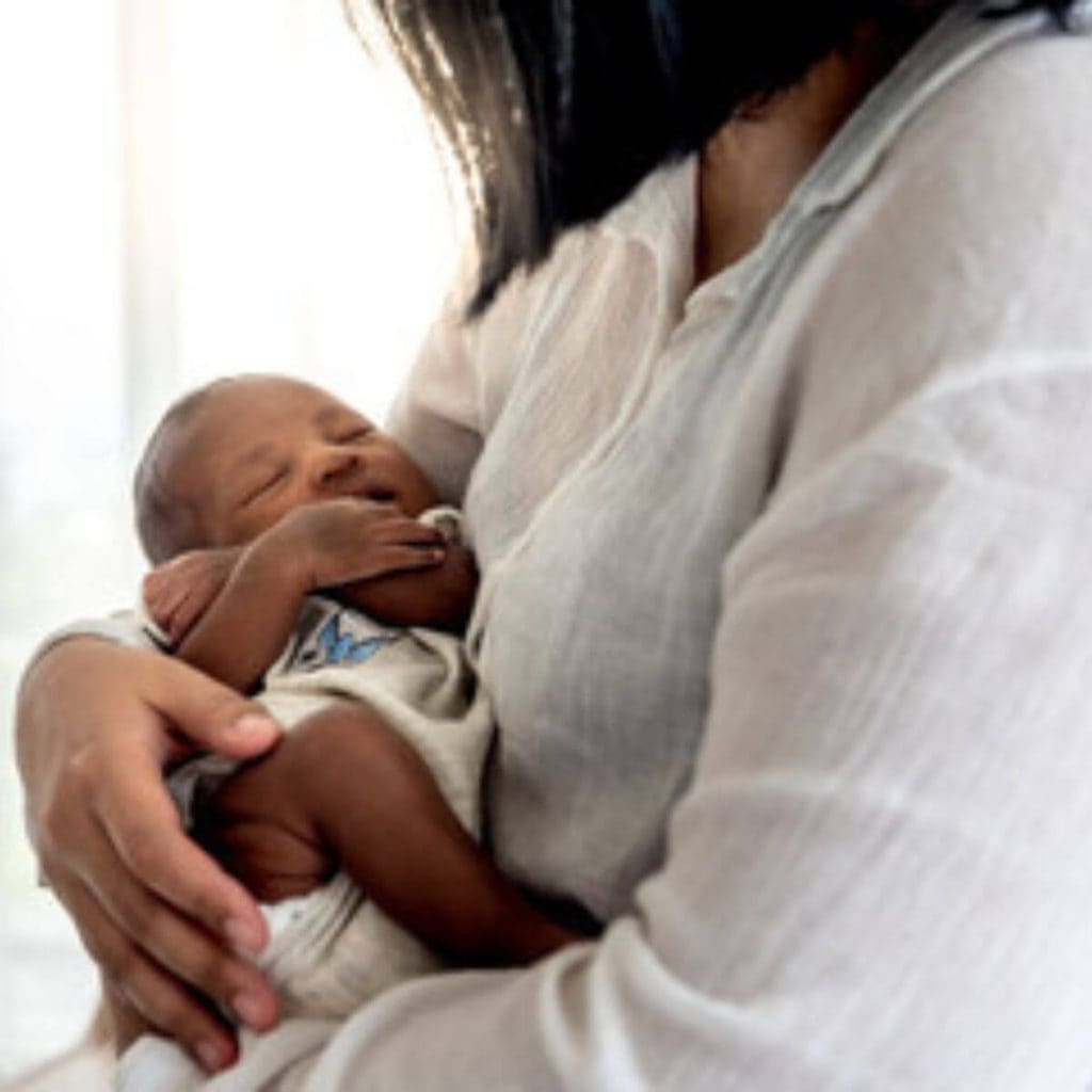 An African American woman is standing in front of a window. She is wearing a white shirt and is cradling a newborn baby in her arms